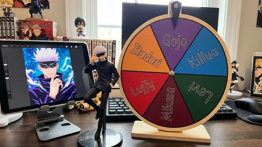 A wheel spinner on a laptop screen showing anime character names as colorful segments including Itadori, Killua, Levi, Mikasa, Luffy, and Gojo in bright colors with the result arrow pointing to Gojo, placed on a desk beside an anime figure and a tablet showing an anime episode
