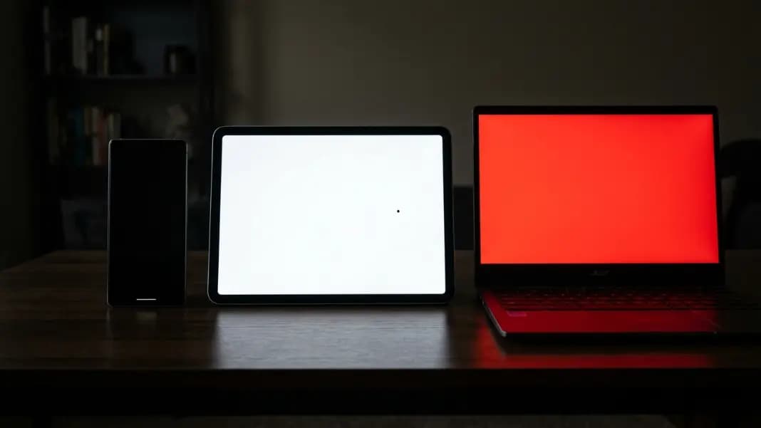 A phone, tablet, and Chromebook displayed side by side each showing the dead pixel test running in fullscreen mode, the phone with a black background, the tablet with a white background showing a small black dot, and the Chromebook with a red background, all devices at maximum brightness against a dark neutral background