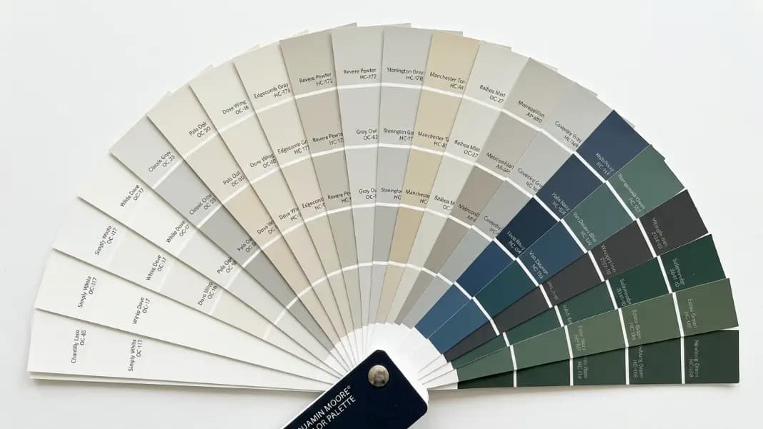 A flat lay of paint color fan deck strips arranged in an arc showing color progressions from light neutrals to warm grays to deep blues and greens, with color name labels visible on each chip, photographed from directly above on a white surface