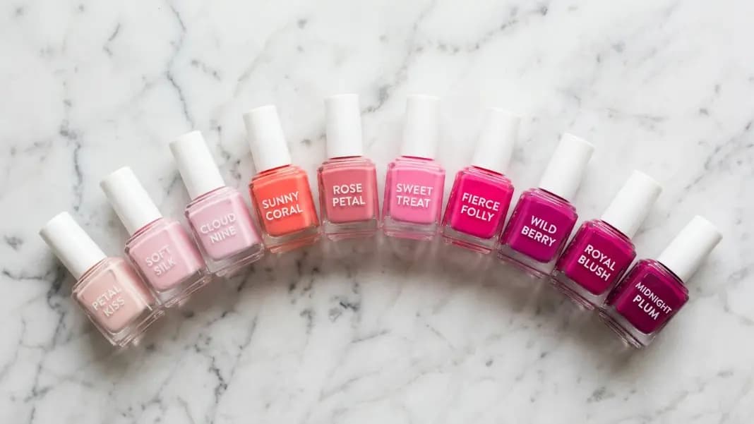 A flat lay of pink nail polish bottles and swatches arranged in a gradient from pale blush and baby pink on the left through coral, rose, and hot pink in the middle to deep fuchsia and magenta on the right, each labeled with its color name and hex code, on a white marble surface A flat lay of pink nail polish bottles and swatches arranged in a gradient from pale blush and baby pink on the left through coral, rose, and hot pink in the middle to deep fuchsia and magenta on the right, each labeled with its color name and hex code, on a white marble surface